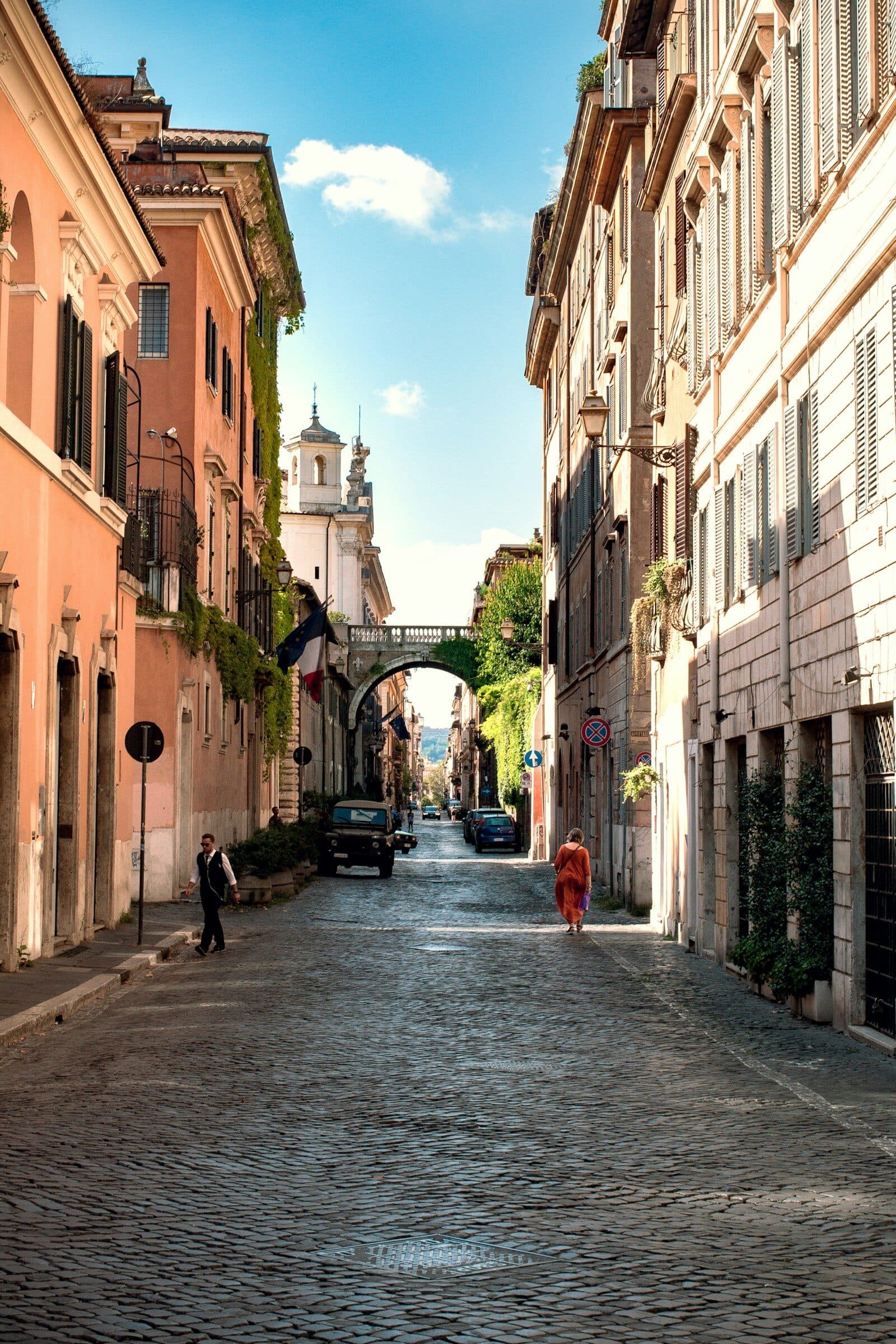 Una via di paese in salita, con un arco in fondo e due persone che camminano al sole.