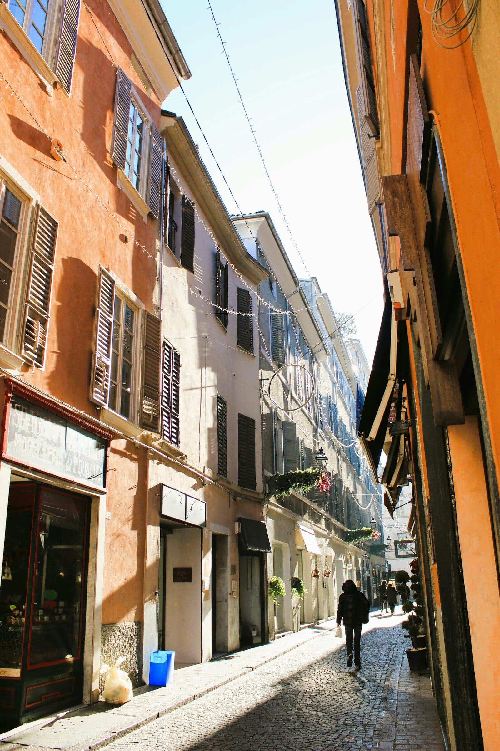 Una persona cammina da sola in una via stretta, tra palazzi alti, con il sole che arriva dal fondo.